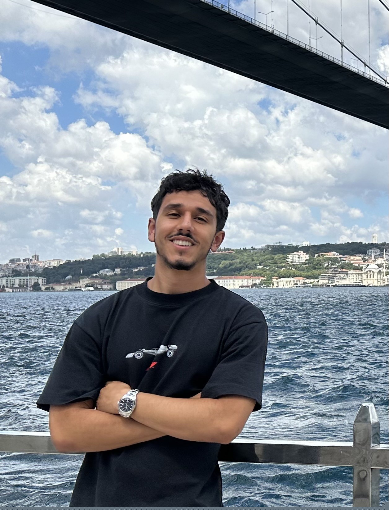 A young tan man with short, dark curly hair and facial hair wraing a black graphic t-shirt crosses his arms and smiles at the camera in front of a lake background.